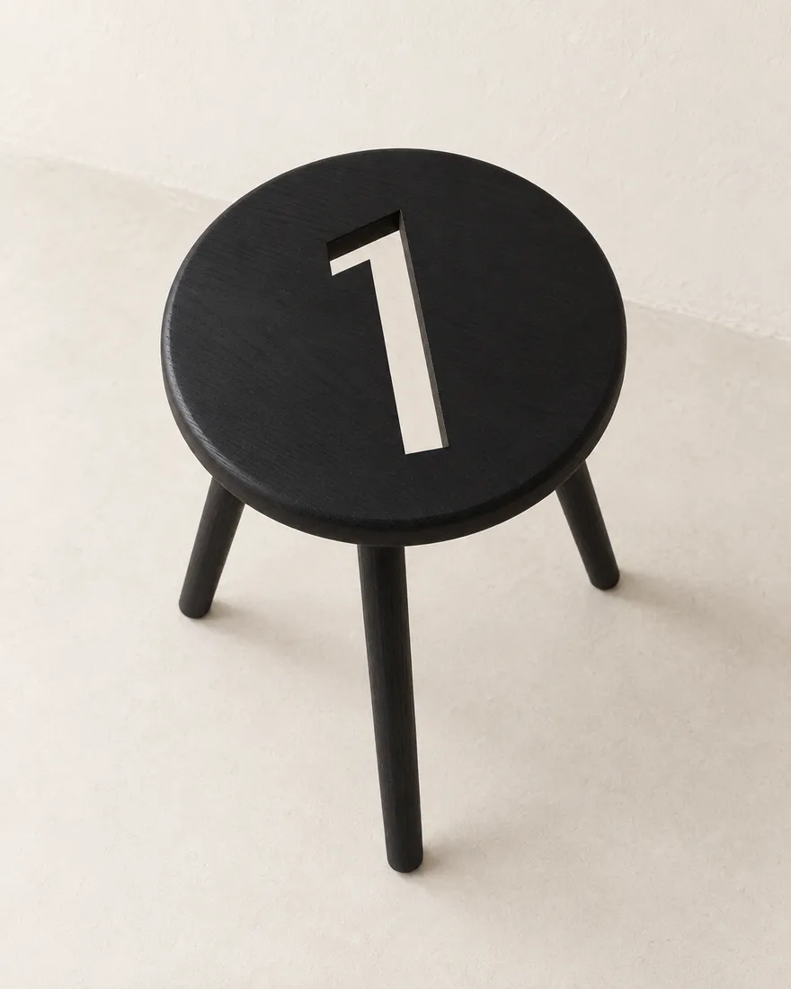 A small round black stool on three slim legs, the leaning 1 cut clean through its seat.