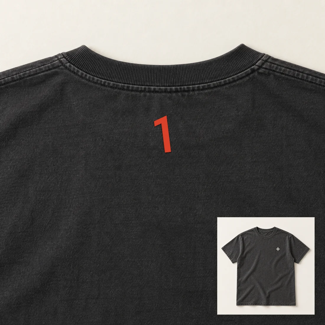 Close on the back of a washed black tee: a vermilion 1 stitched just below the collar. Inset, the front: a small bone plus on the chest.